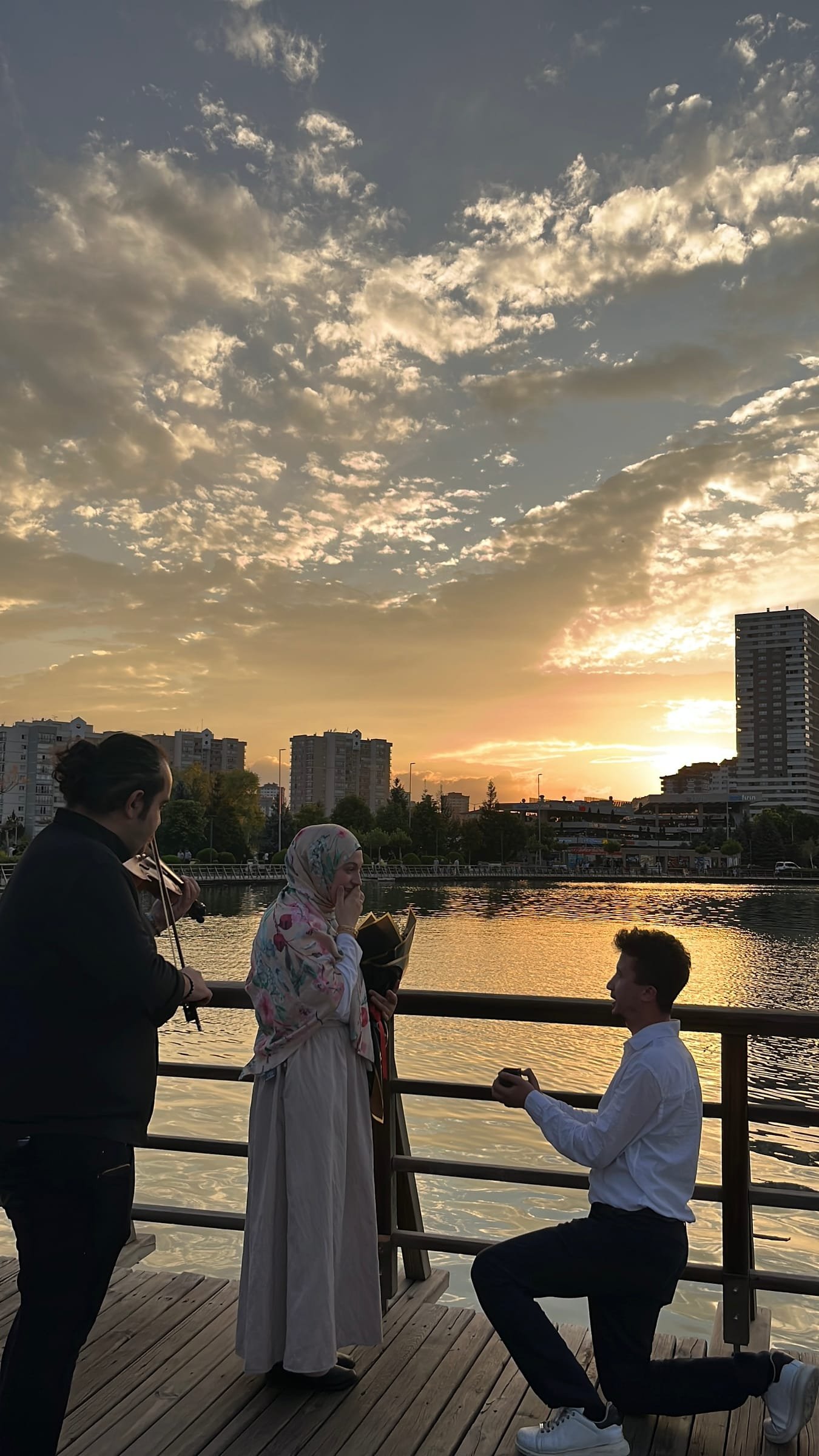 Gün batımında göl kenarındaki iskelede diz çökerek evlilik teklifi eden damat adayı ve keman çalan müzisyen