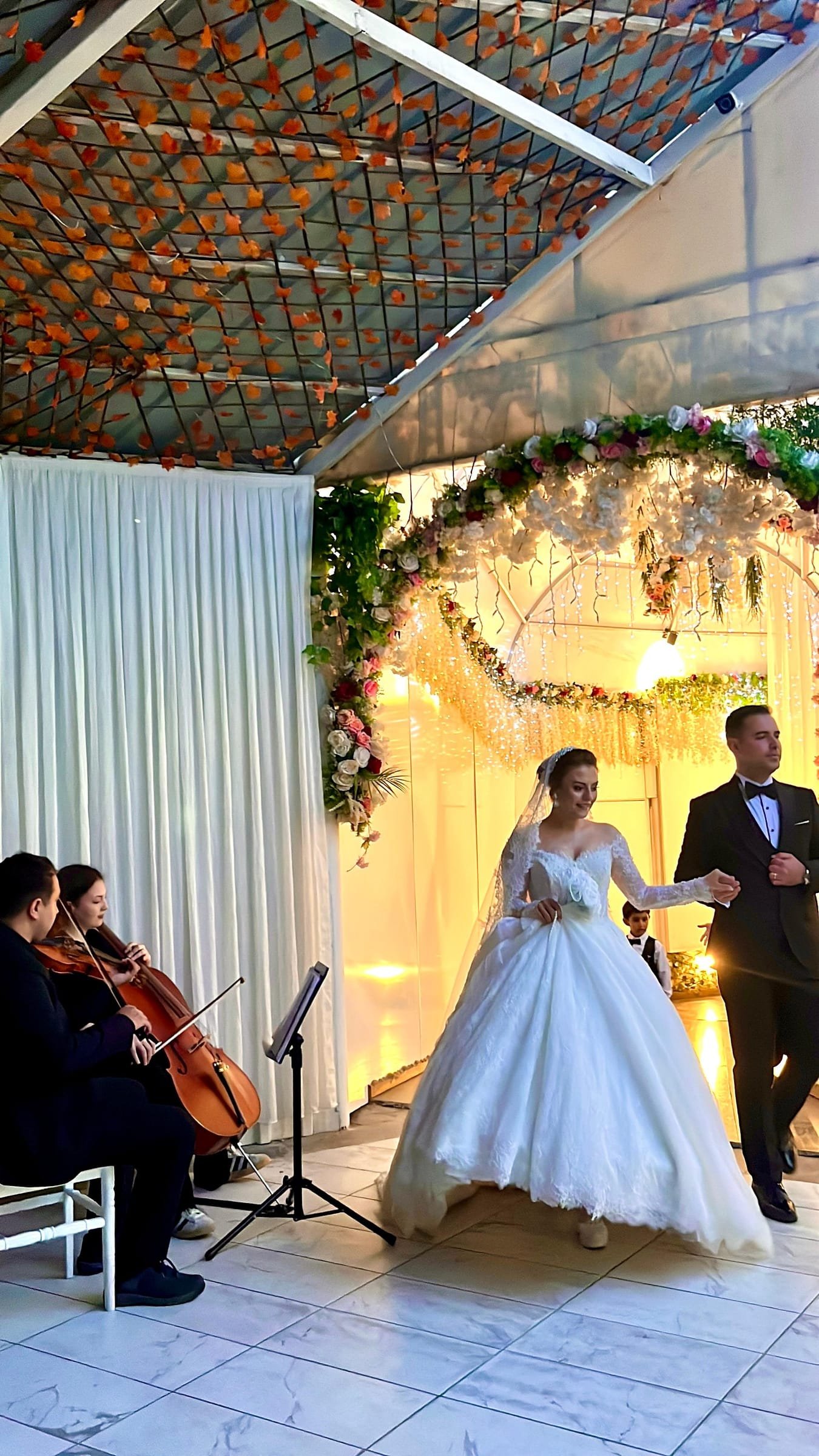 Keman ve çello ikilisi eşliğinde çiçek kemerin altından giren gelin ve damat