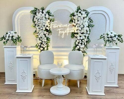 Beyaz çiçekli arka plan, dekoratif sehpalar ve puflarla hazırlanmış nişan konsepti